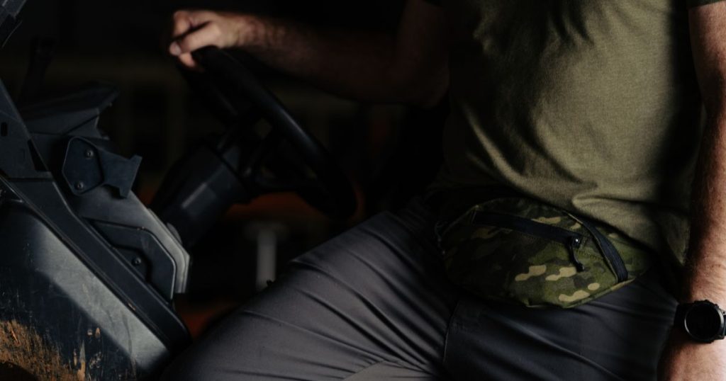 A man sitting in a cart wearing a Blue Alpha CCW Fanny Pack in camo green.
