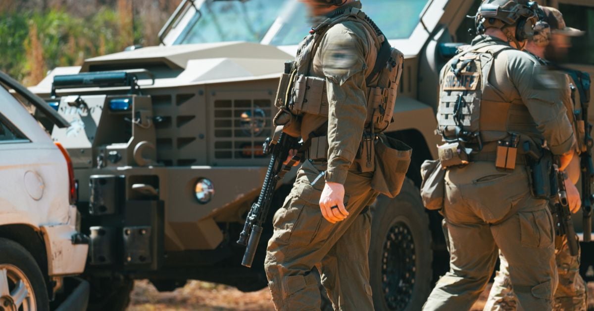 Several men in full plate carrier gear and carrying shooting gear in a field.